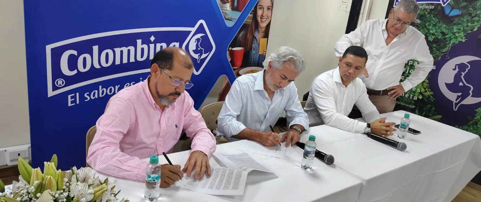 Tres hombres sentados, uno de ellos firma un documento. Al fondo un banner de la empresa Colombina