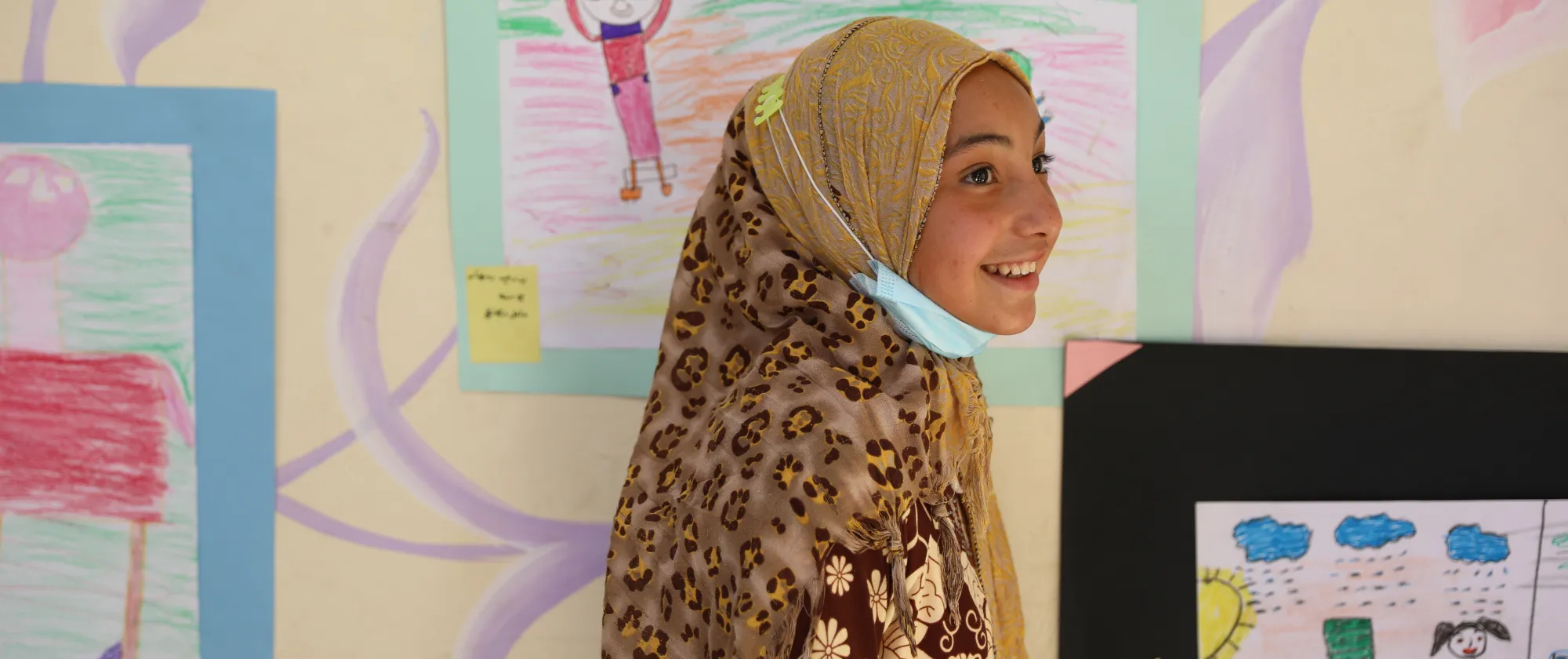 Young girl smiling in front of colourful children's artwork, wearing a patterned headscarf and a mask around her neck.