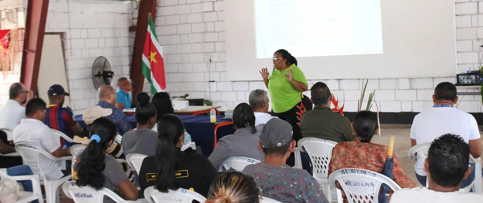 A woman in a green shirt presents to a seated audience in a room with the flag of Suriname.