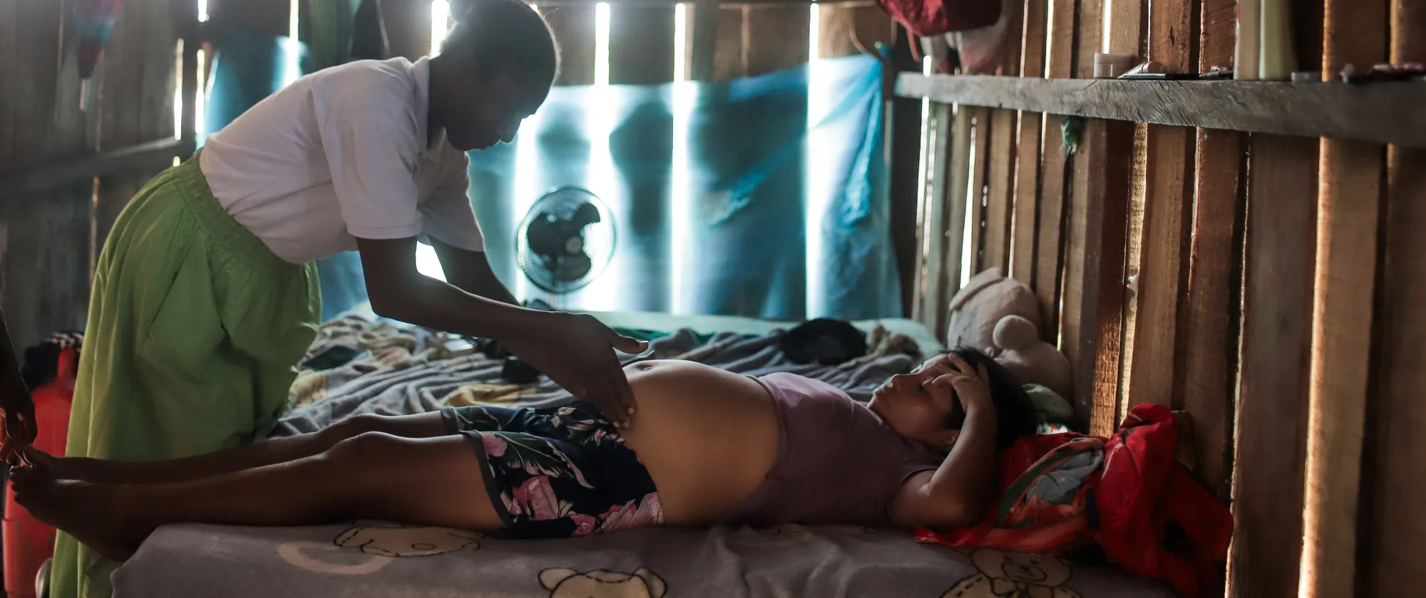 Mujer afrodescendiente tocando el abdomen de una mujer indígena embarazada recostada en una cama dentro de una vivienda con paredes de madera. Un ventilador y telas colgadas son visibles en el fondo rústico.