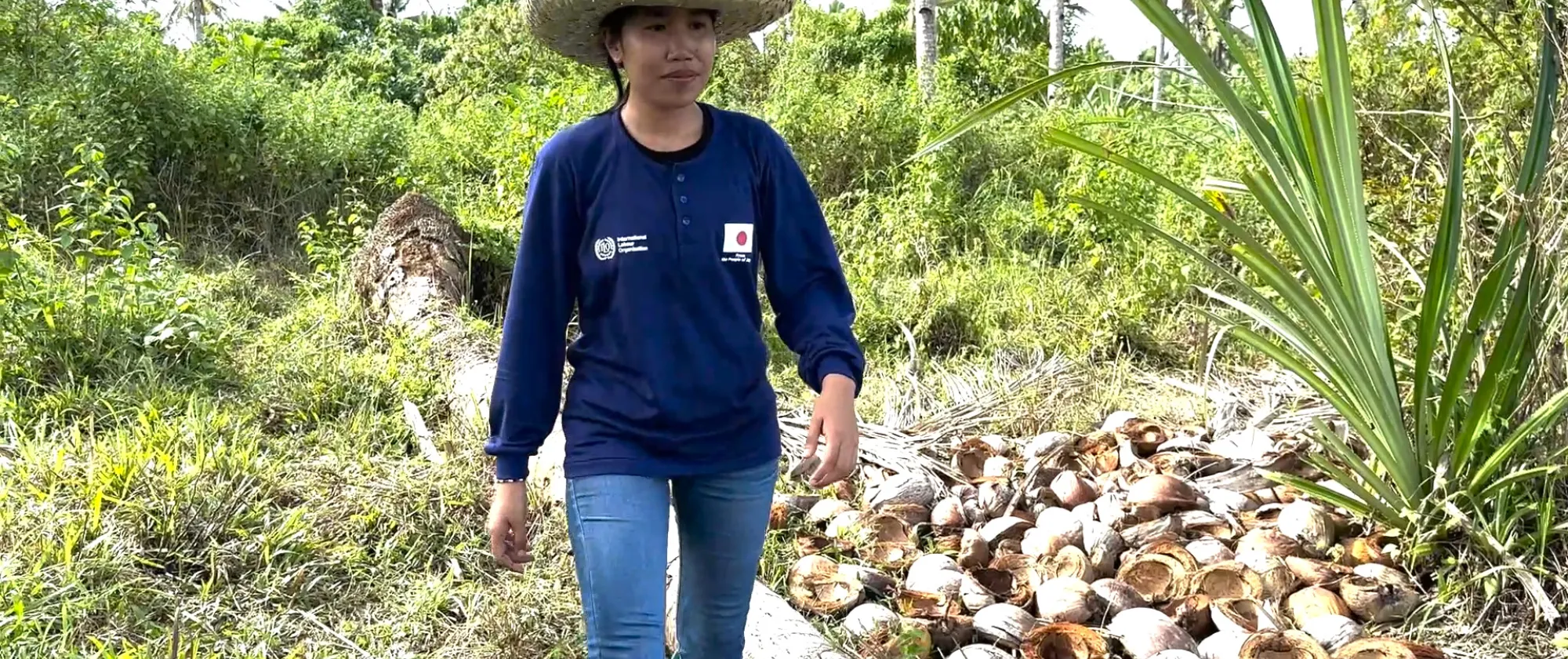 a worker wearing long-sleeve short and hat walks along a coconut farm with fallen trees