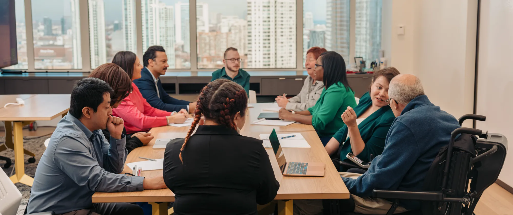 Several people sitting around a table with laptops and paper in front of them. A large window is behind them with tall buildings outside.