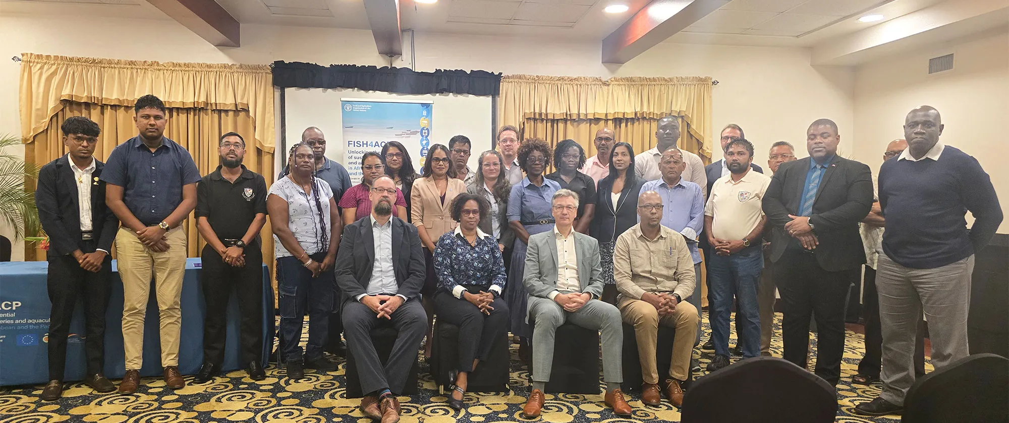Group photo of approximately 30 participants standing and seated indoors at a professional event. Behind them is a banner displaying the logo of the FAO and the FISH4ACP project. 