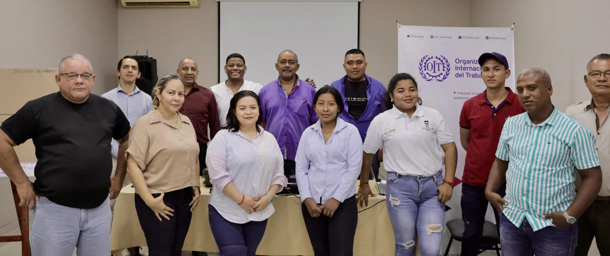 Grupo de 13 personas, 4 mujeres y 9 hombres, posando sonrientes. Detrás el pendón de la OIT.