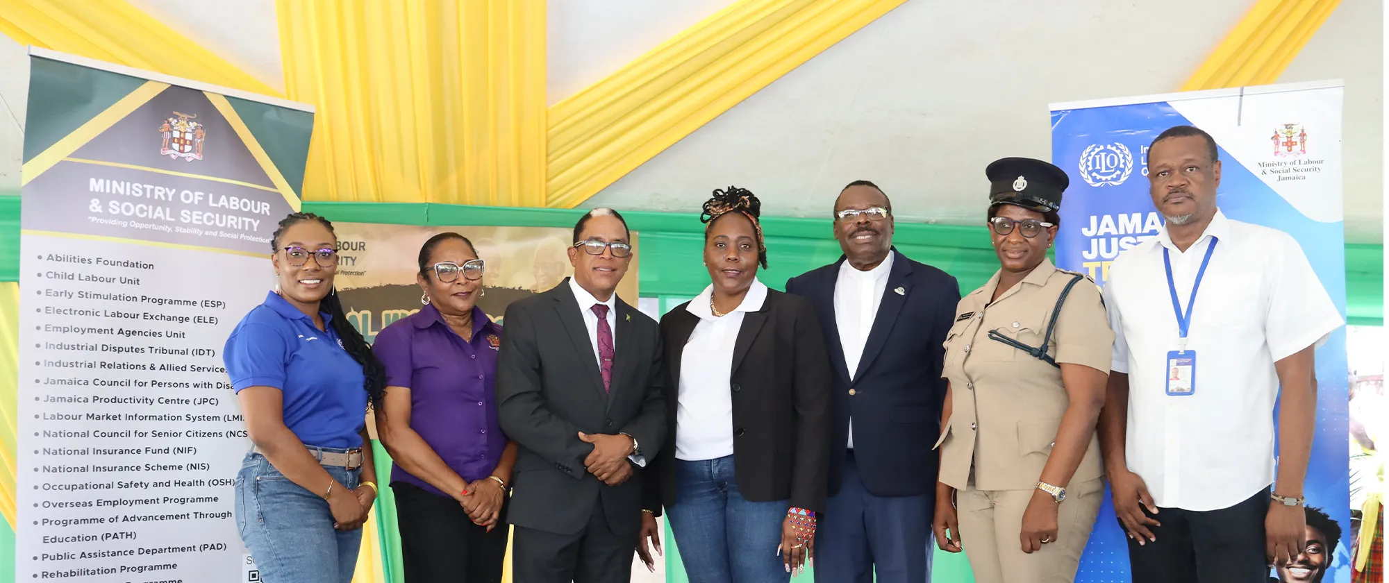 A group of seven individuals stands together in front of banners for the Ministry of Labour & Social Security and the International Labour Organization at an indoor event. 