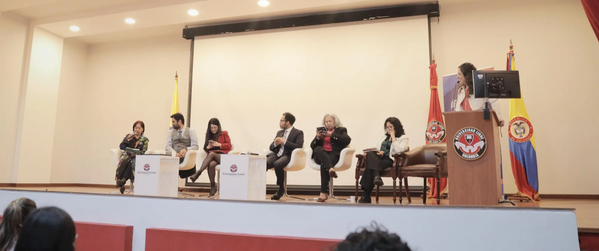 Tarima de un auditorio con 5 personas sentadas en frente y una mujer en un atril, de fondo se vislumbra la bandera de Colombia y de la Universidad Libre