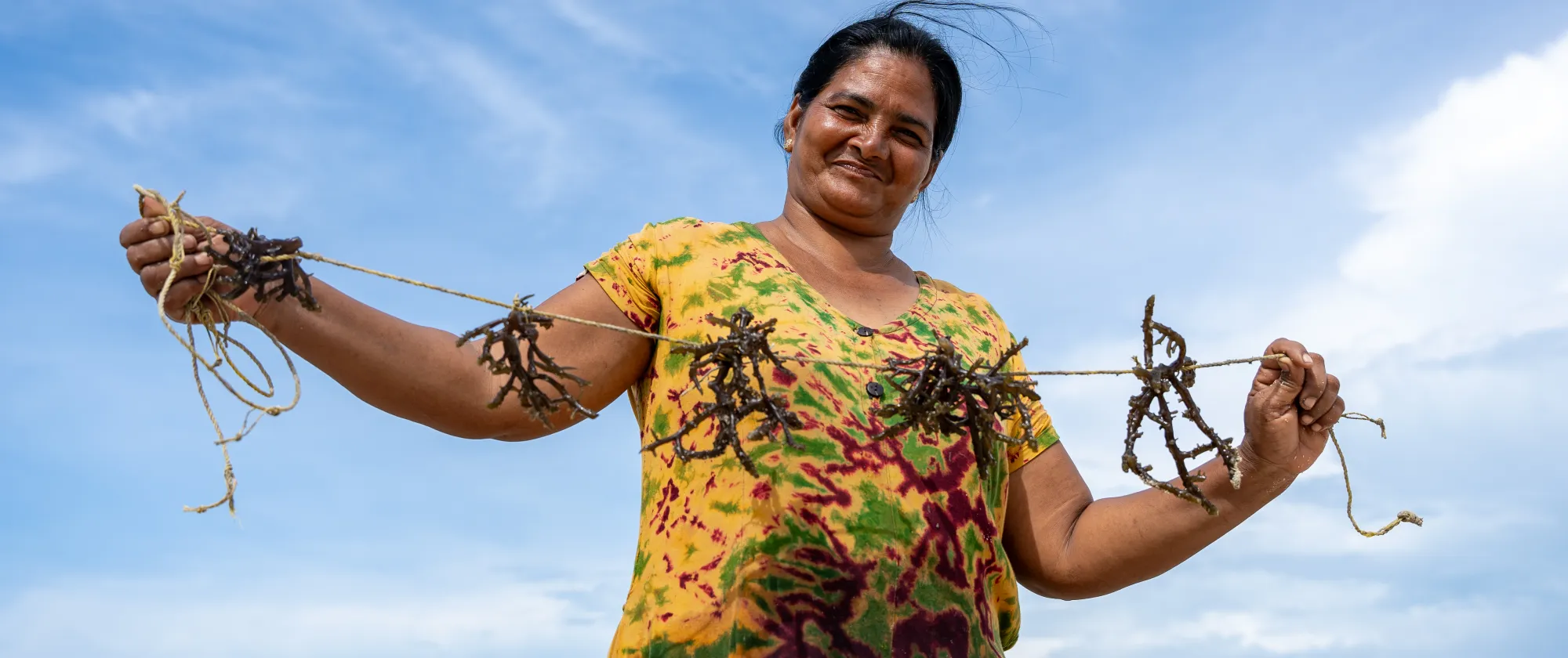A woman stands against the vast blue sky, she holdes a rope with seaweed tied to it for cultivation