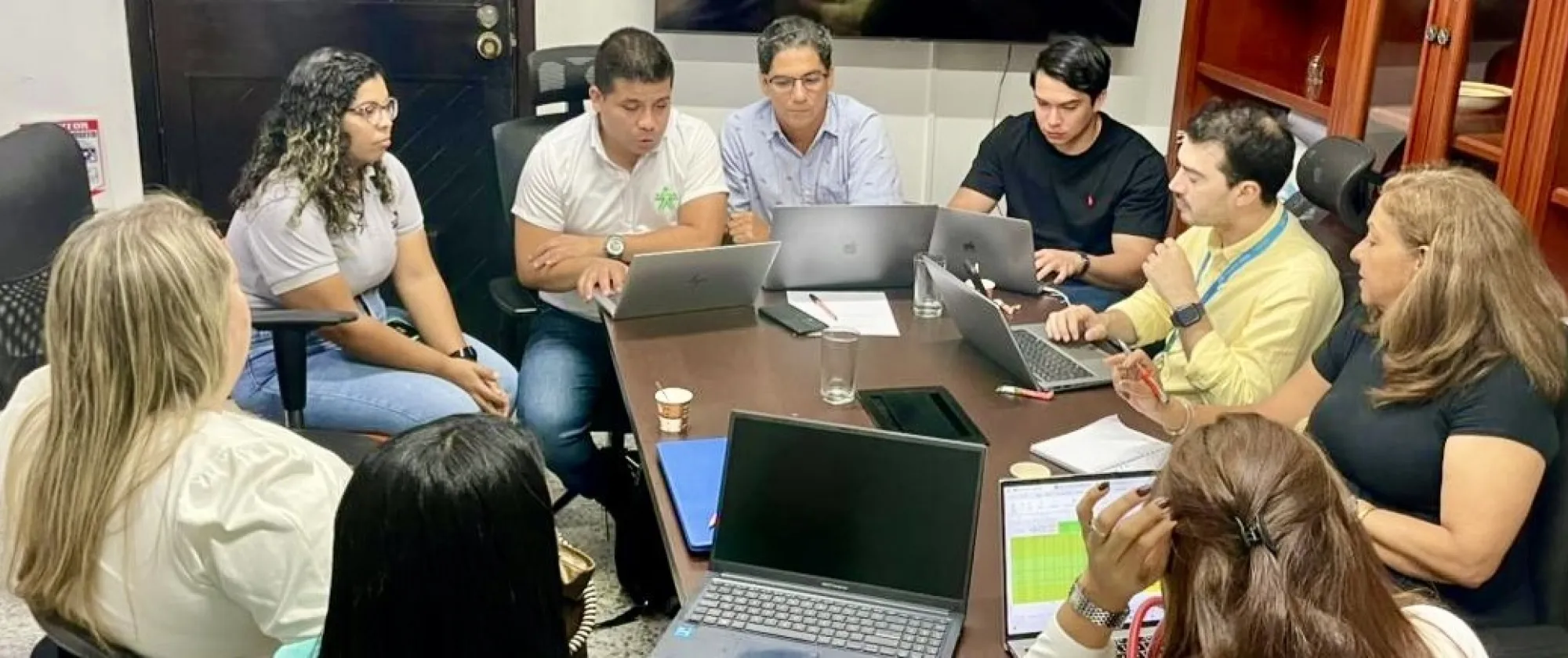 Nueve personas están sentadas alrededor de una mesa rectangular, todas enfocadas en sus portátiles o tomando notas. Se trata de un grupo diverso de hombres y mujeres, con edades aproximadas entre los 25 y 50 años. La mayoría viste ropa informal o semiformal.