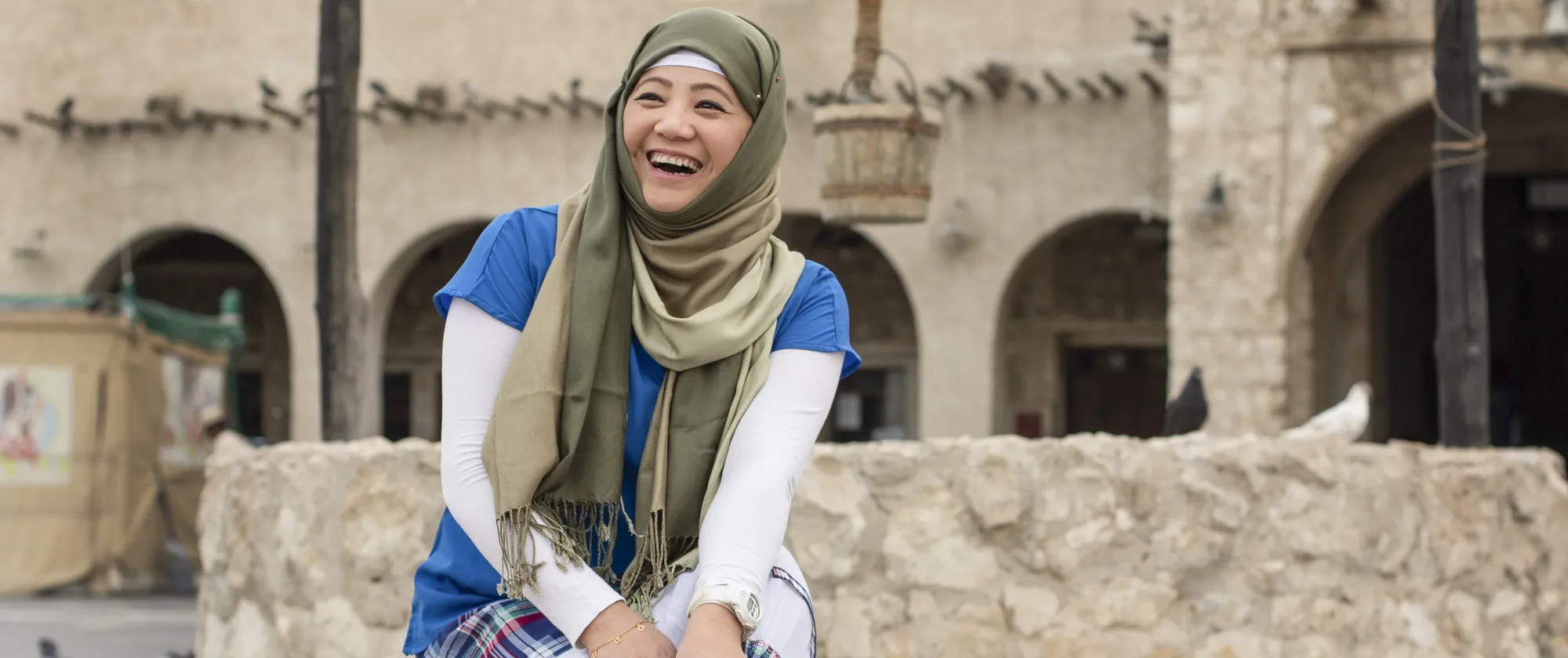 The image shows a person wearing a green headscarf, blue shirt, and white long sleeves sitting on a stone wall in front of an old building with arched doorways and pigeons perched on its ledges.