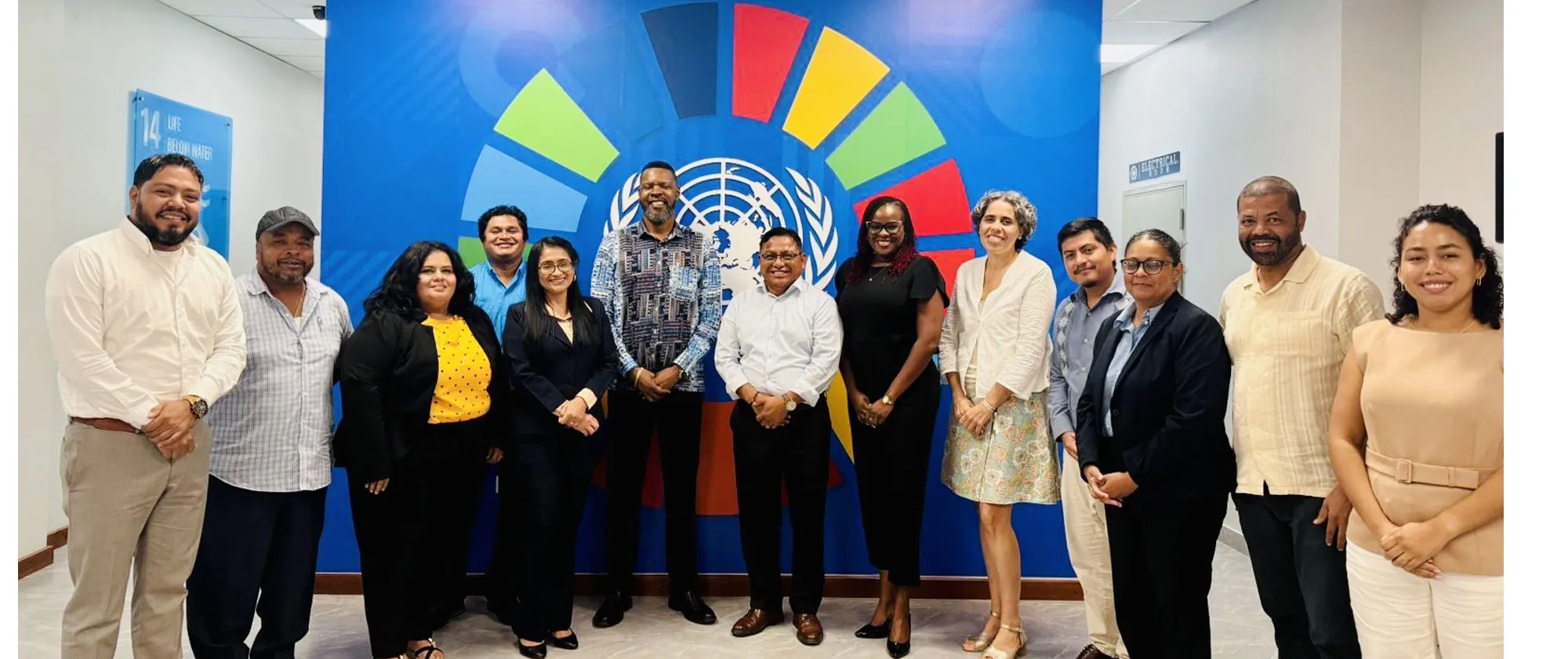 A group of thirteen people stands together indoors in front of a large blue wall featuring the United Nations emblem and colorful segments forming a circular design. 