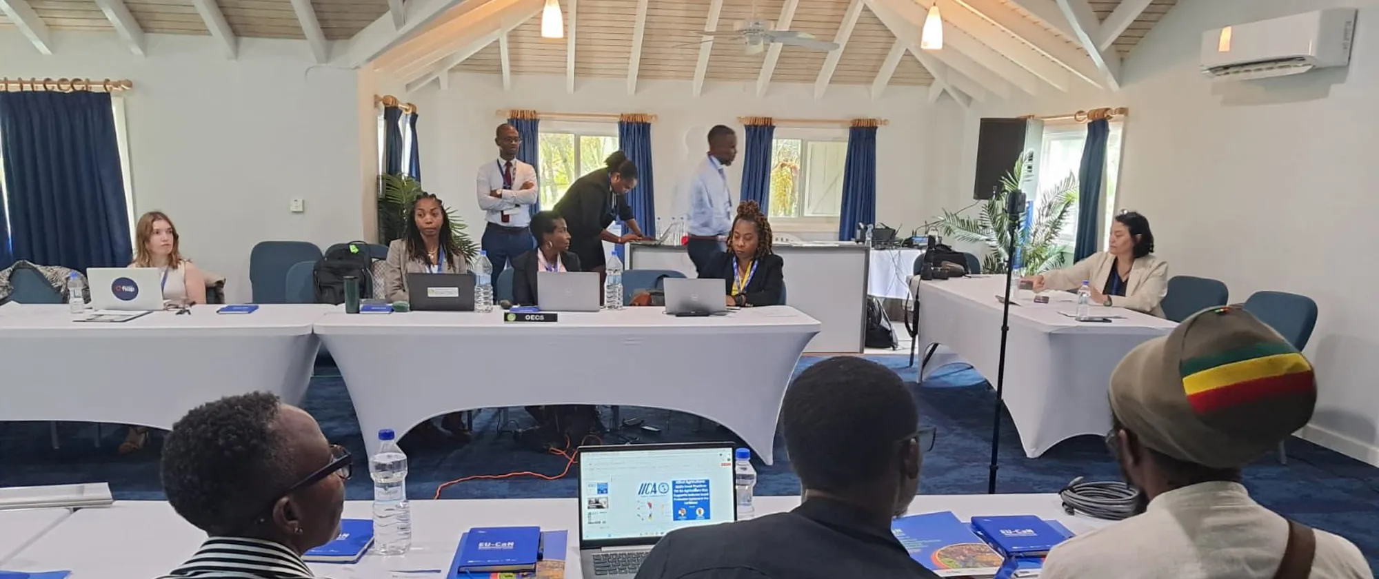 A group of people is participating in a  workshop with attendees are seated around U-shaped tables, working on laptops and engaging in discussion