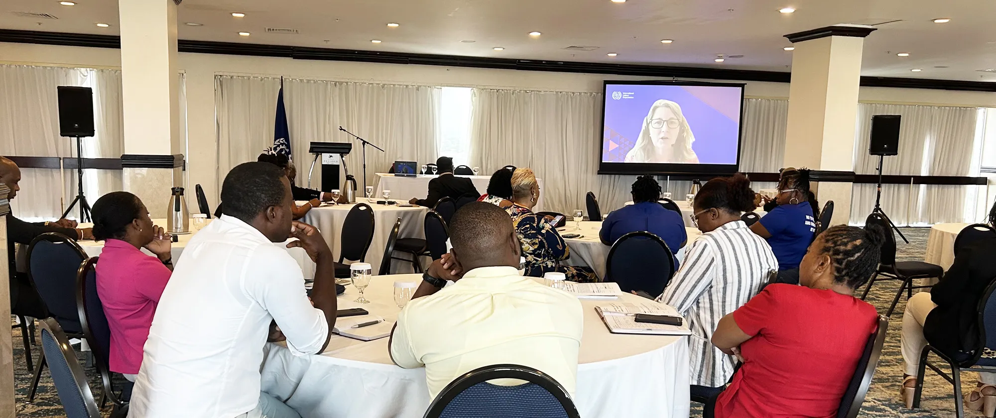 A group of people is seated at round tables in a conference room, attentively watching a presentation projected onto a large screen at the front of the room.