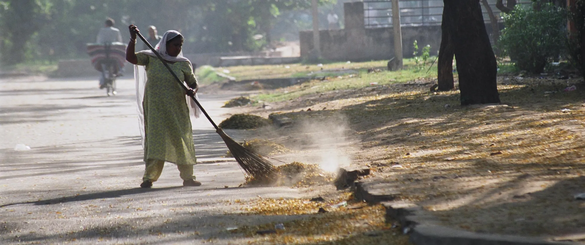 The image shows an Indian woman sweeping leaves and debris off a tree-lined street with a broom, while another person rides a bicycle in the background.