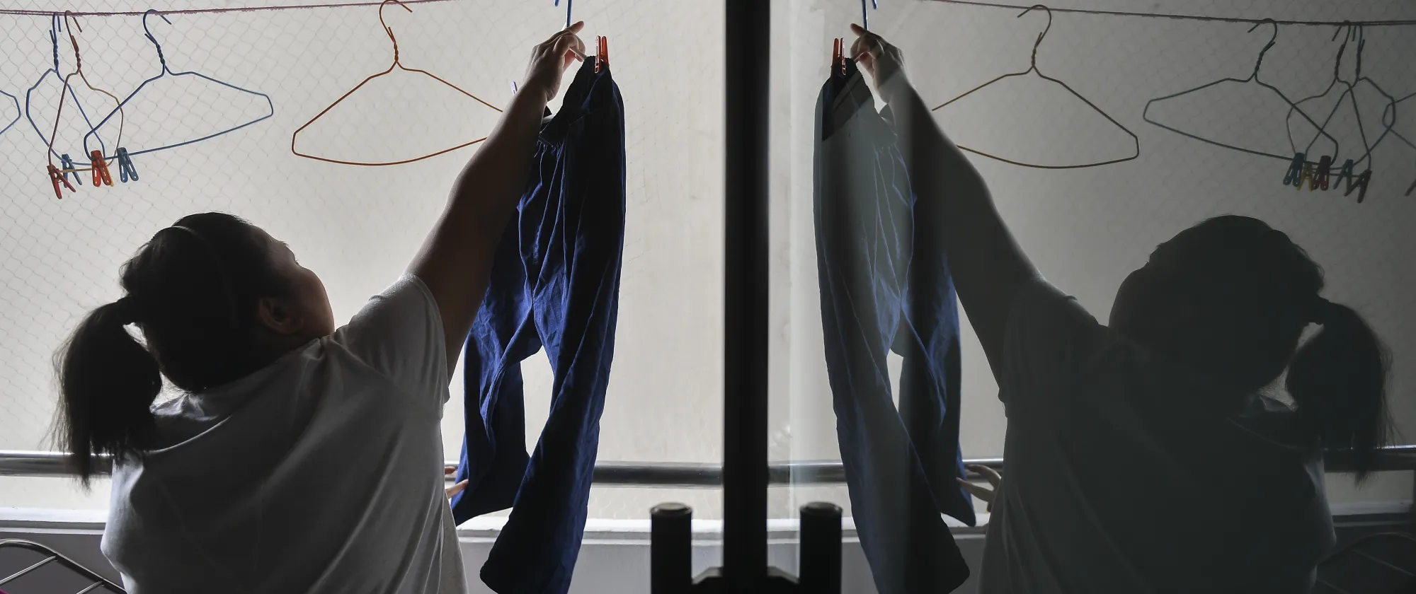 The image shows a woman hanging blue pants on a clothesline with several empty hangers, as the scene is reflected in a glass window.