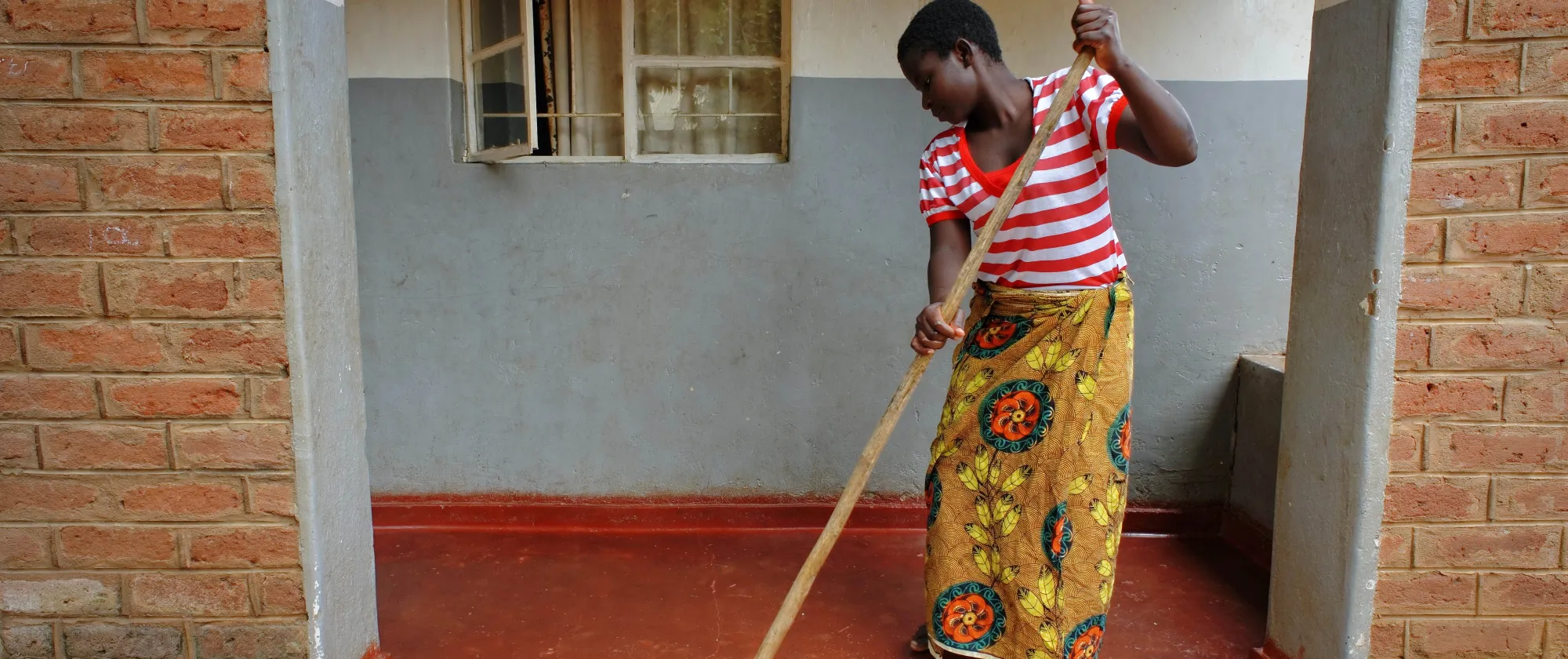 The image shows an African woman sweeping the floor of a porch area with a broom, positioned between two brick walls and in front of a windowed wall.