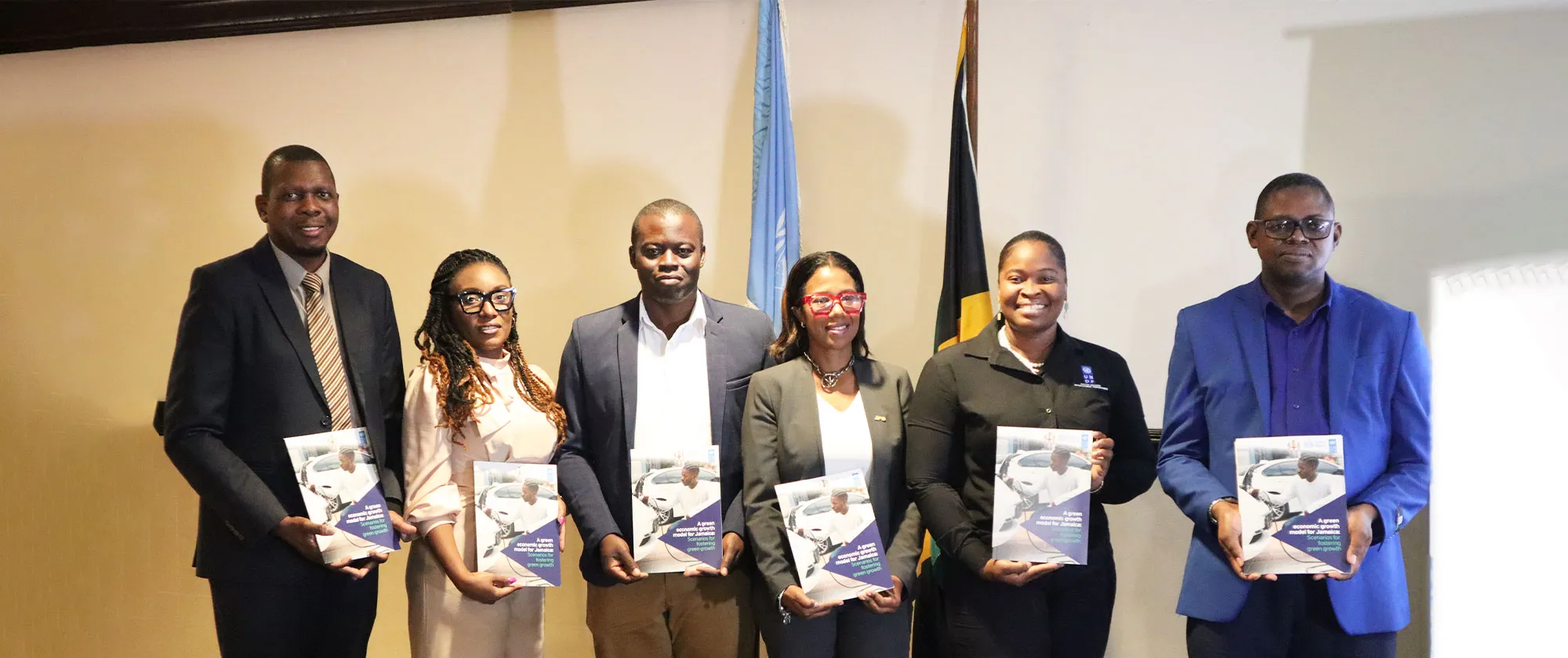 Six individuals are standing in a row indoors, each holding a publication titled "A green economic growth model for Jamaica: Towards green growth." 