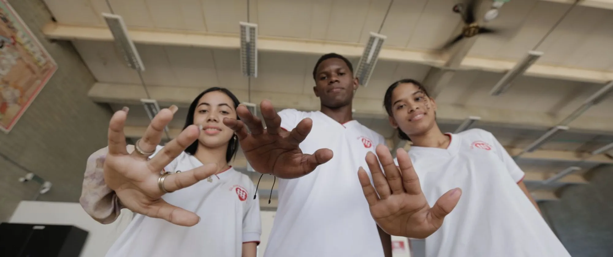 En la imagen aparecen tres jóvenes, dos mujeres a los lados y un hombre en el centro, están de pie en un espacio escolar. Todos visten camisetas blancas tipo uniforme con un pequeño logo rojo en el pecho. 