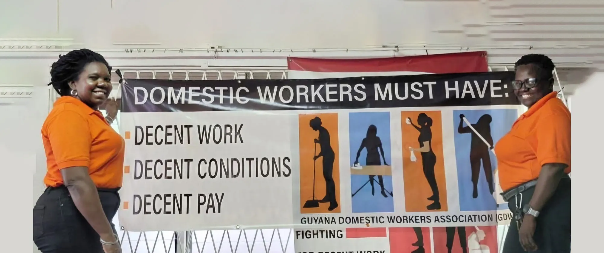 Two women, both wearing orange shirts and standing side by side, are positioned in front of a large banner advocating for the rights of domestic workers. The banner reads "DOMESTIC WORKERS MUST HAVE: DECENT WORK, DECENT CONDITIONS, DECENT PAY"
