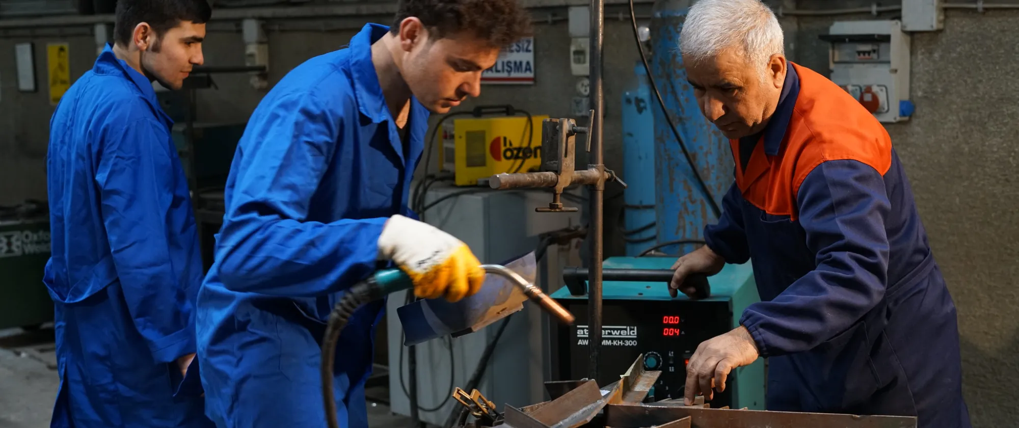 young man handling a soldering iron next to another young man and under the supervision of an older man