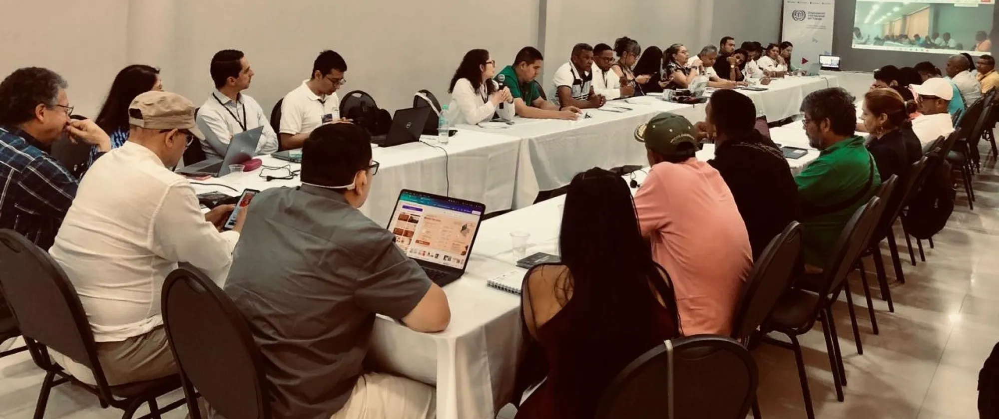 Reunión con más de 30 personas sentadas en forma de U alrededor de mesas largas, algunas con computadoras portátiles. Al fondo, una mujer habla con micrófono. Se proyecta una imagen y al lado un pendón de la OIT