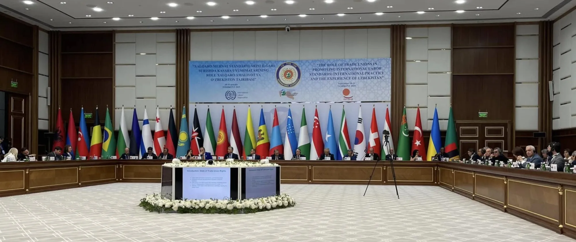 A group of people sitting in a big room with flags in front of a wall