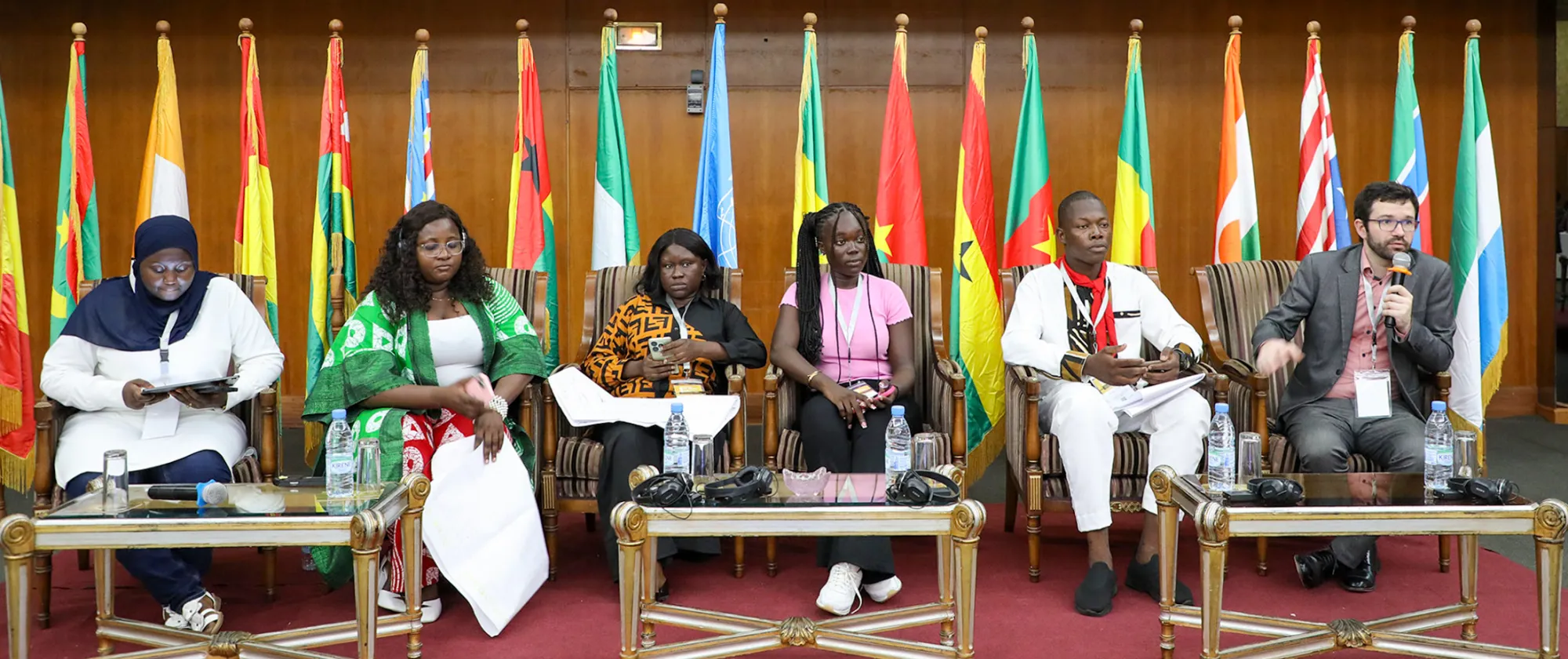 Youth delegate speaks into a microphone during a panel discussion at the 4th Annual West Africa and Sahel Youth Forum in Dakar, seated alongside fellow participants with country flags in the background.
