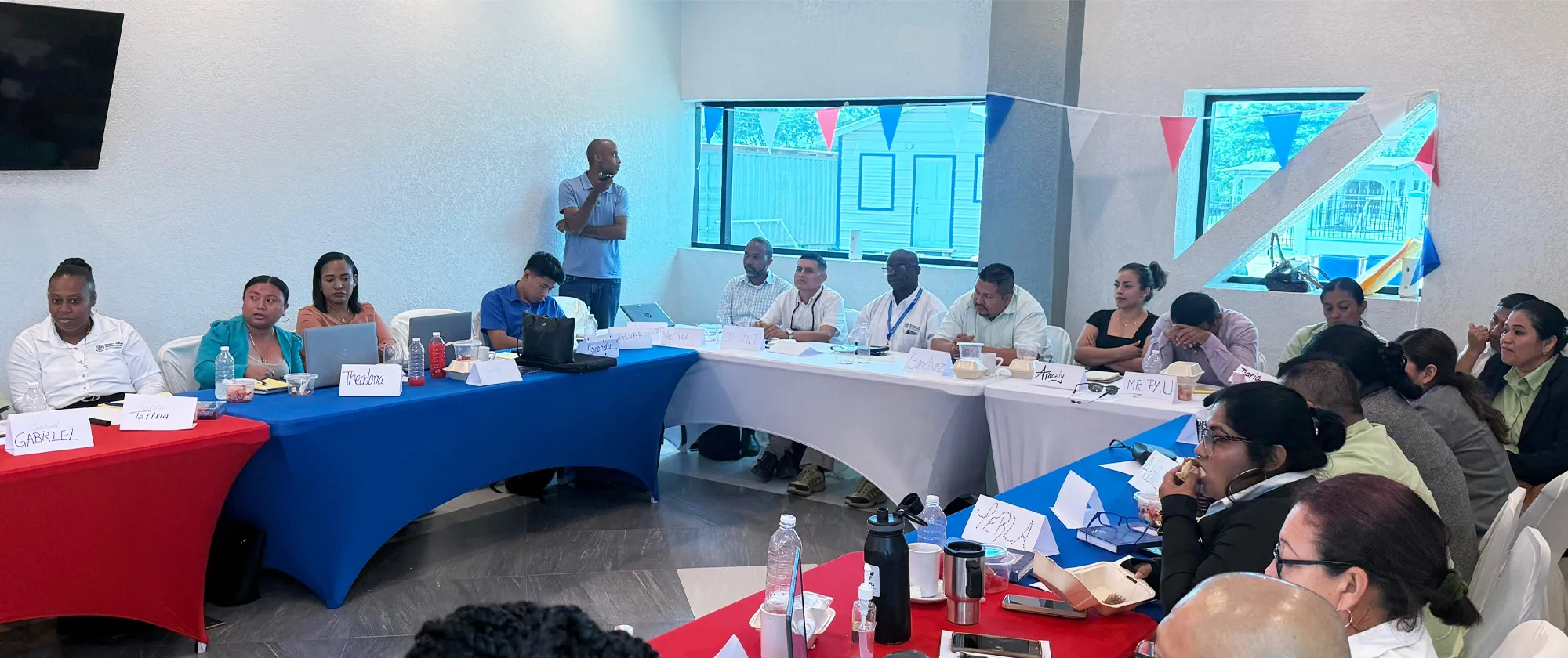 A group of people are seated at long tables arranged in a U-shape in a brightly lit room listening to Abdelmalik Muhummed, ILO Specialist, Employment and Labour Market speak on public employment services in Belize.