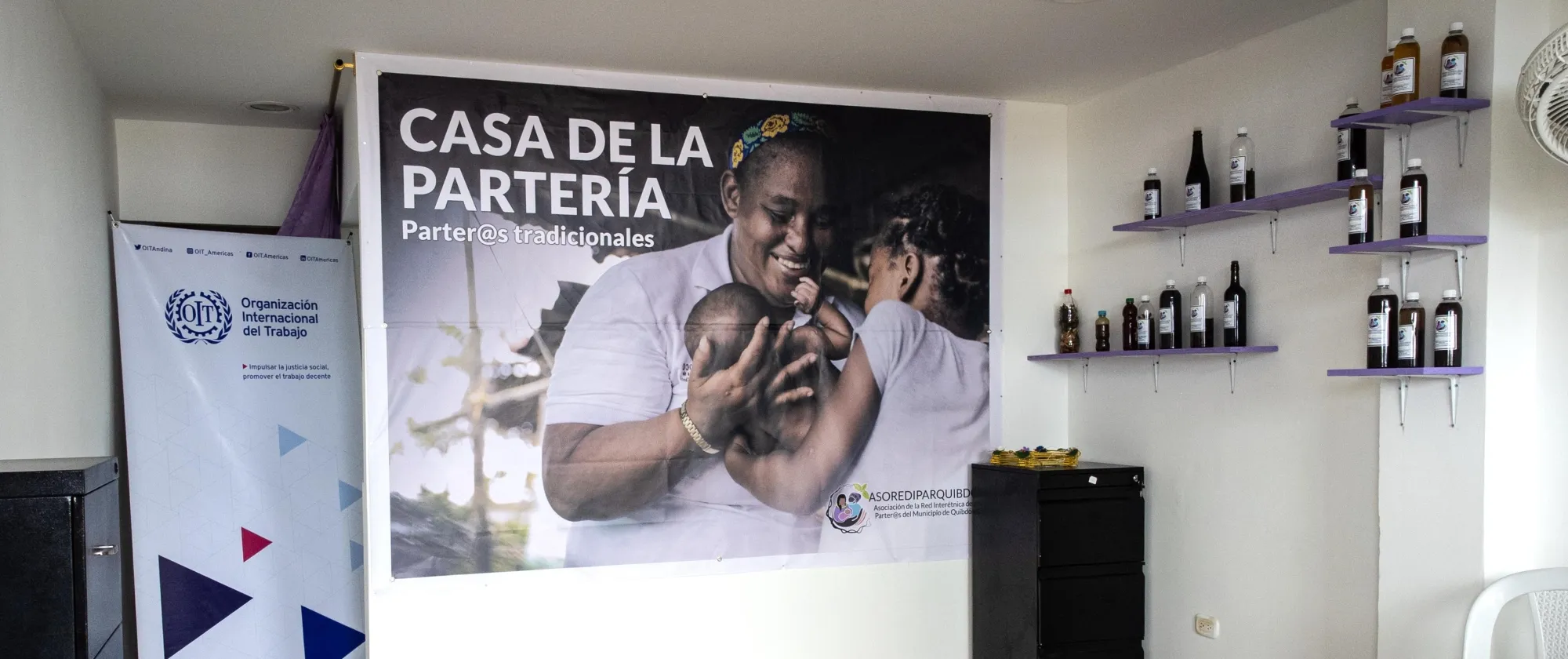Interior de la nueva Casa de la Partería en Quibdó. En la pared se ve un gran afiche con la imagen de una partera afrodescendiente sonriendo mientras carga a un bebé, acompañado del texto “Casa de la Partería – Parter@s tradicionales” y el logo de ASOREDIPAR. A la izquierda hay un pendón de la OIT y a la derecha estantes con frascos de preparados medicinales.