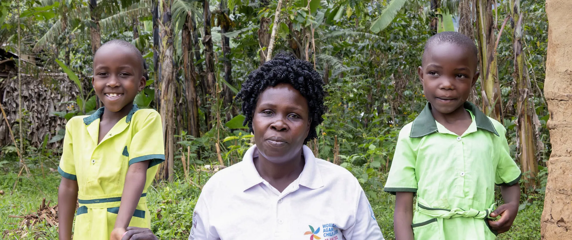 A woman and her two young children stand outside their community kindergarten in rural eastern Uganda, surrounded by green banana trees.