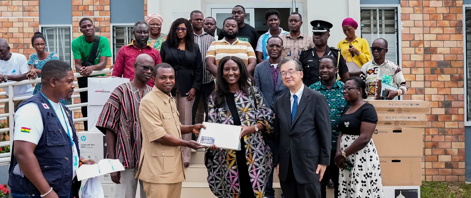 The Japanese Ambassador to Ghana, Hiroshi Yoshimoto, and ILO Country Director Vanessa Phala smiling as they present a laptop to a local representative in Bibiani, Ghana. They are joined by officials and community members, with boxes of donated equipment visible in the background.