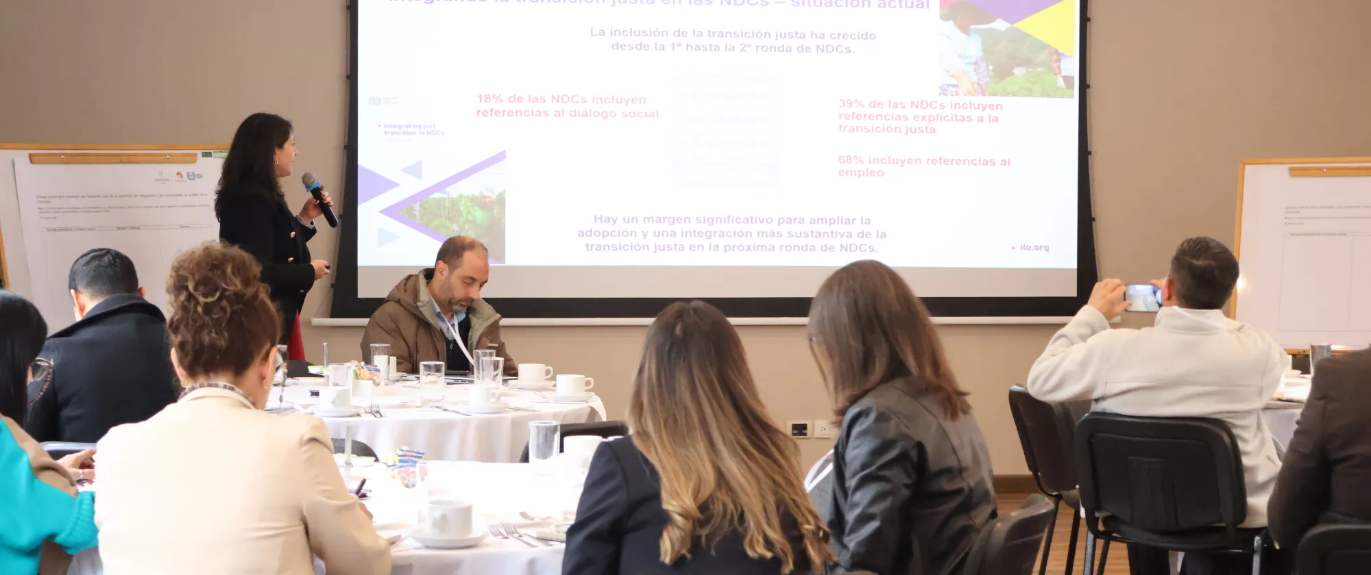 Una mujer realiza una presentación ante un grupo de personas en una sala de conferencias. En la pantalla se muestra una diapositiva titulada “Integrando la transición justa en las NDCs – situación actual”, con datos y porcentajes sobre la inclusión de la transición justa, el diálogo social y el empleo en las NDCs. Los asistentes están sentados en mesas redondas, tomando notas y observando la exposición.