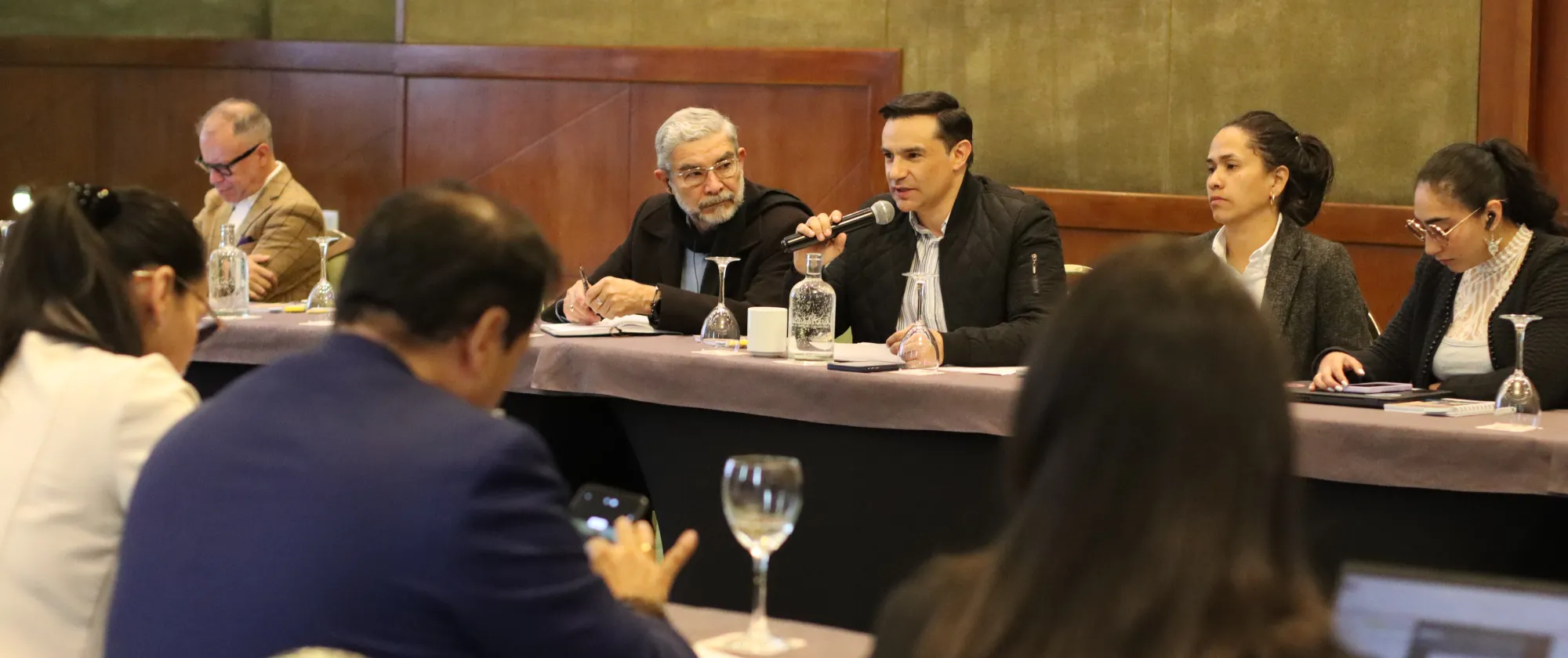 Hombre sostiene un microfono mientras habla a una mesa de reunión de ocho personas. Todos se encuentran sentados.