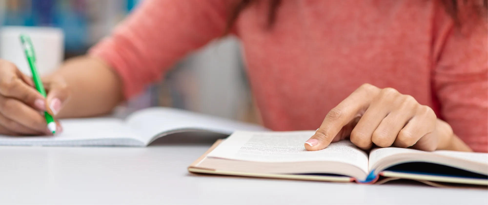 Mujer escribe con lápiz verde en un cuaderno mientras sigue con el dedo las definiciones de un glosario en un libro abierto.