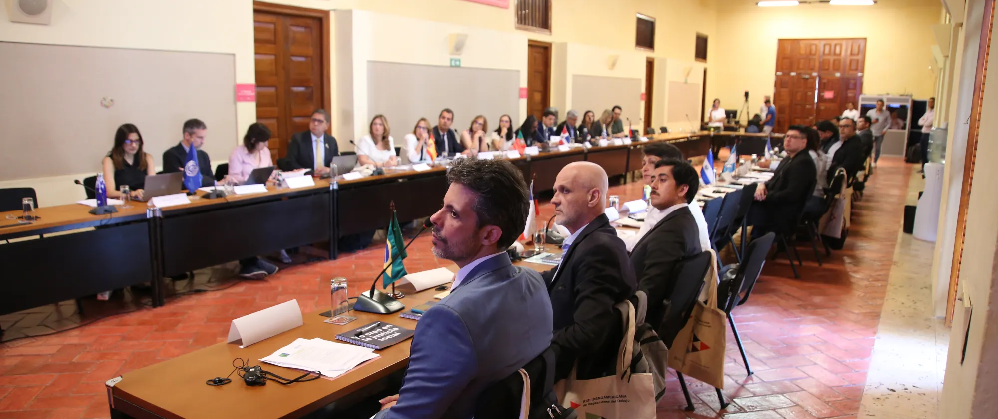 Personas sentadas alrededor de una gran mesa de conferencias en una sala amplia con paredes amarillas y techo alto. Los participantes, vestidos formalmente, escuchan atentamente mientras usan computadoras portátiles y tienen banderas de distintos países frente a ellos.