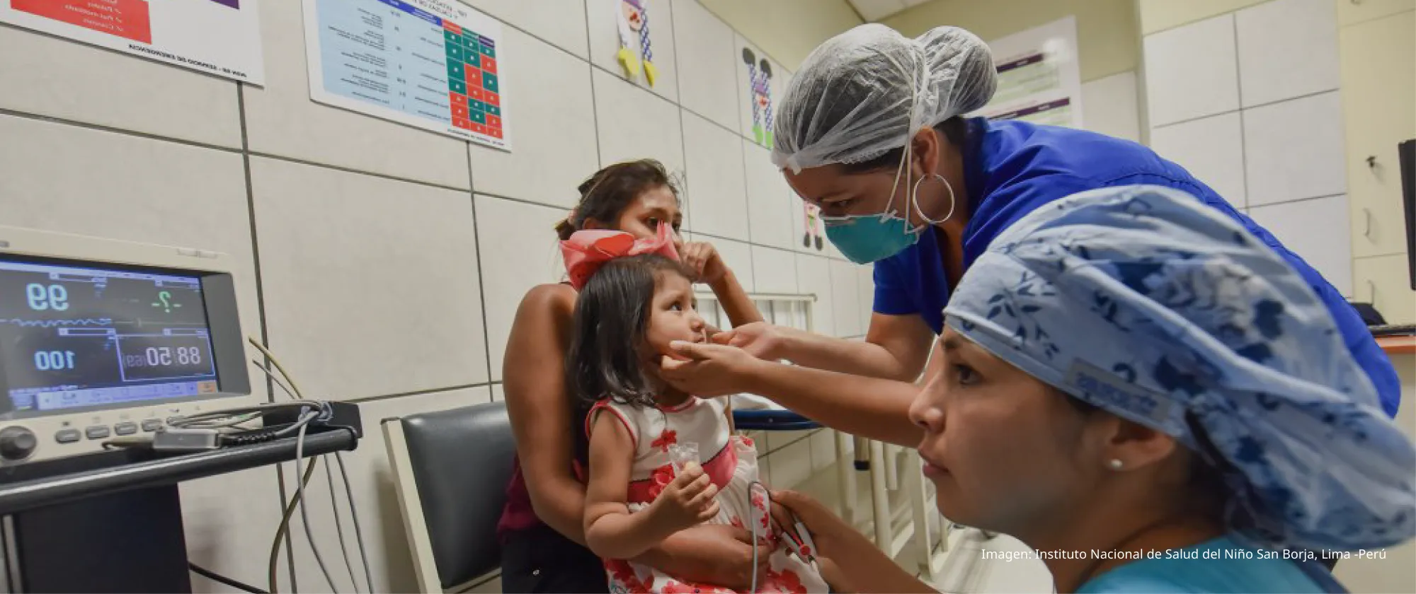 Niña pequeña en vestido rojo sentada en el regazo de su madre mientras una trabajadora de salud le revisa la boca en un consultorio pediátrico.