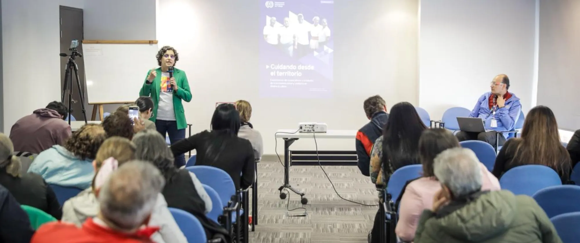 Una persona está de pie al frente de una sala dando una presentación a un grupo de asistentes. Viste un saco verde y sostiene un micrófono. Detrás de ella se proyecta una diapositiva con el título “Cuidando desde el territorio”. El público, compuesto por hombres y mujeres, está sentado en filas de sillas azules, escuchando atentamente.