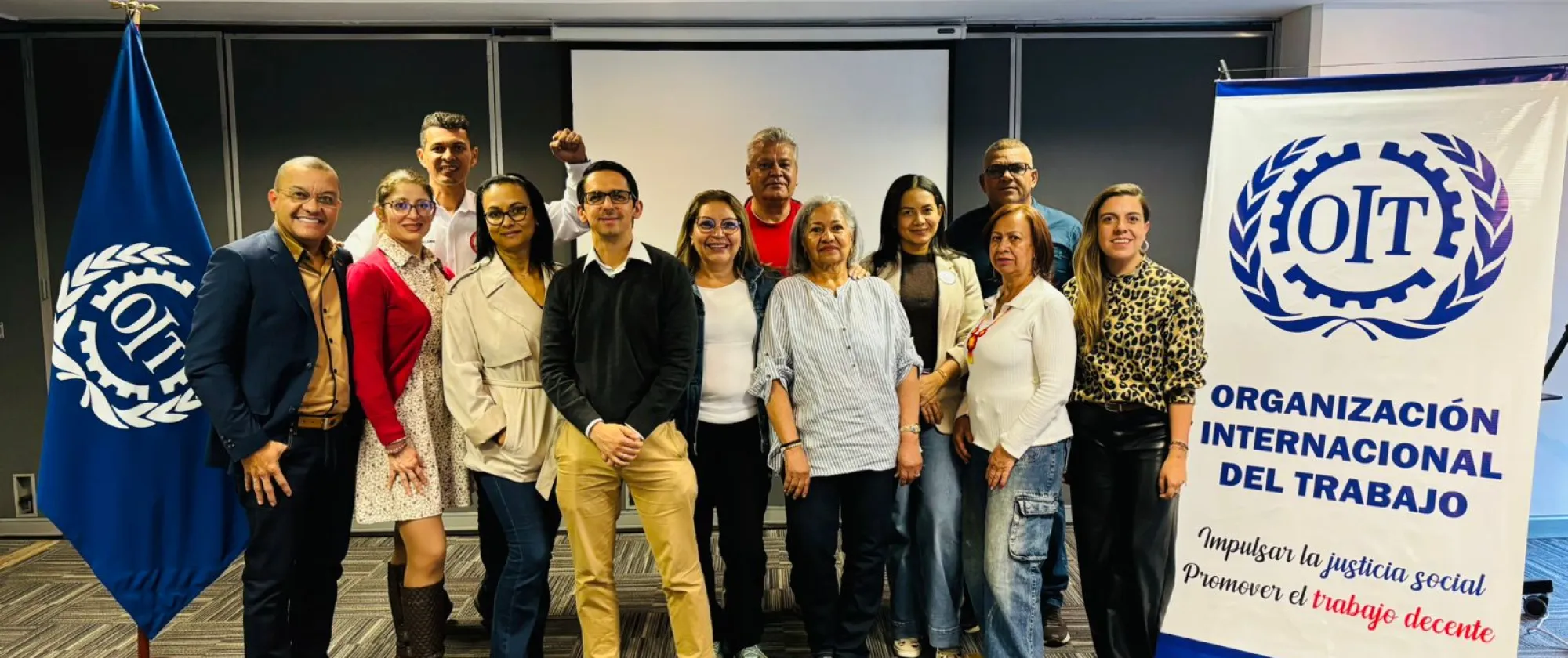 Grupo de doce personas posando en una sala de conferencias junto a una bandera y un cartel de la Organización Internacional del Trabajo (OIT). El cartel muestra el logotipo de la OIT y el lema ‘Impulsar la justicia social. Promover el trabajo decente’. Las personas están sonrientes
