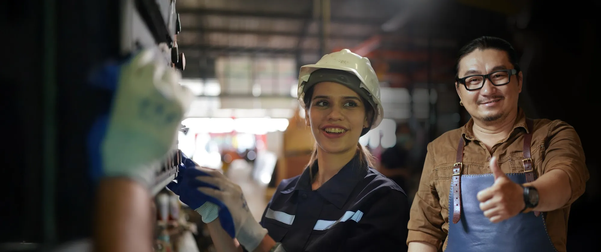 Mujer con casco y overol trabaja en un taller industrial mientras un hombre con gafas y delantal al fondo sonríe y hace señal de pulgar arriba.