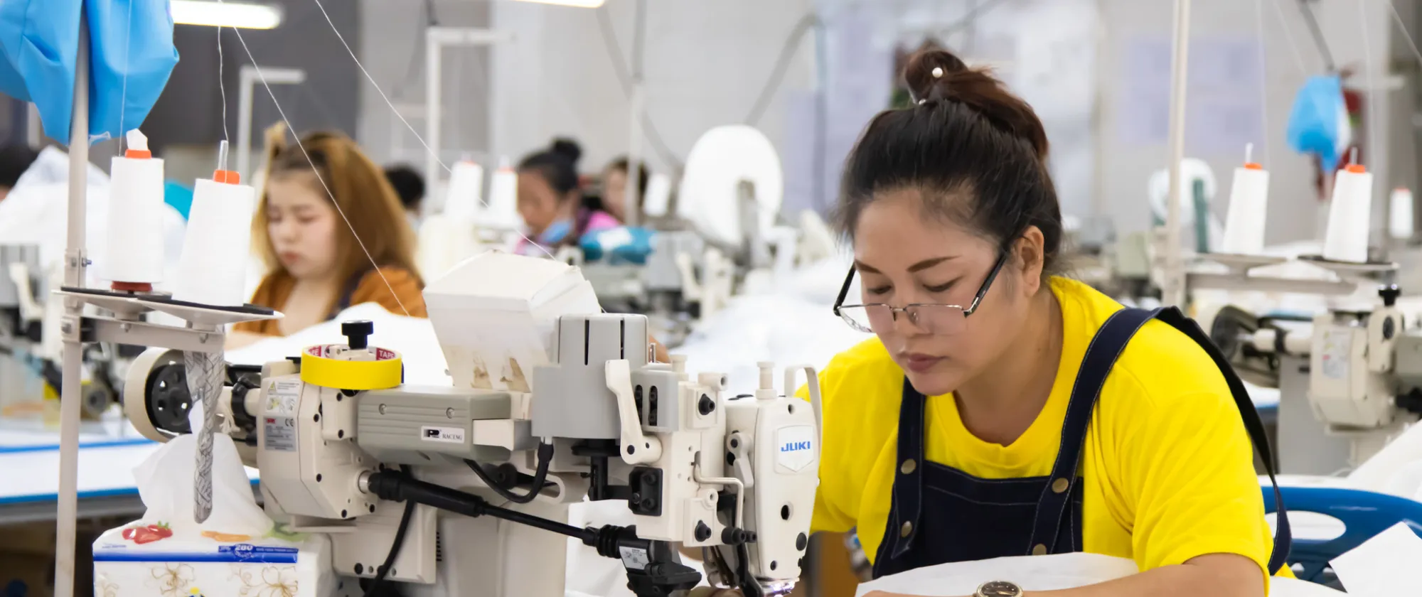 A woman sewing, with sewing machines and other seamstresses working in the background.