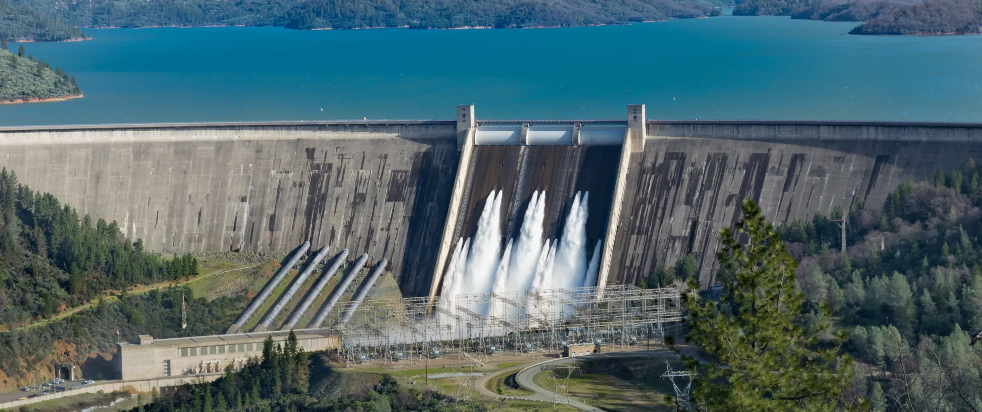 Shasta dam surrounded by roads, trees with lake mountains