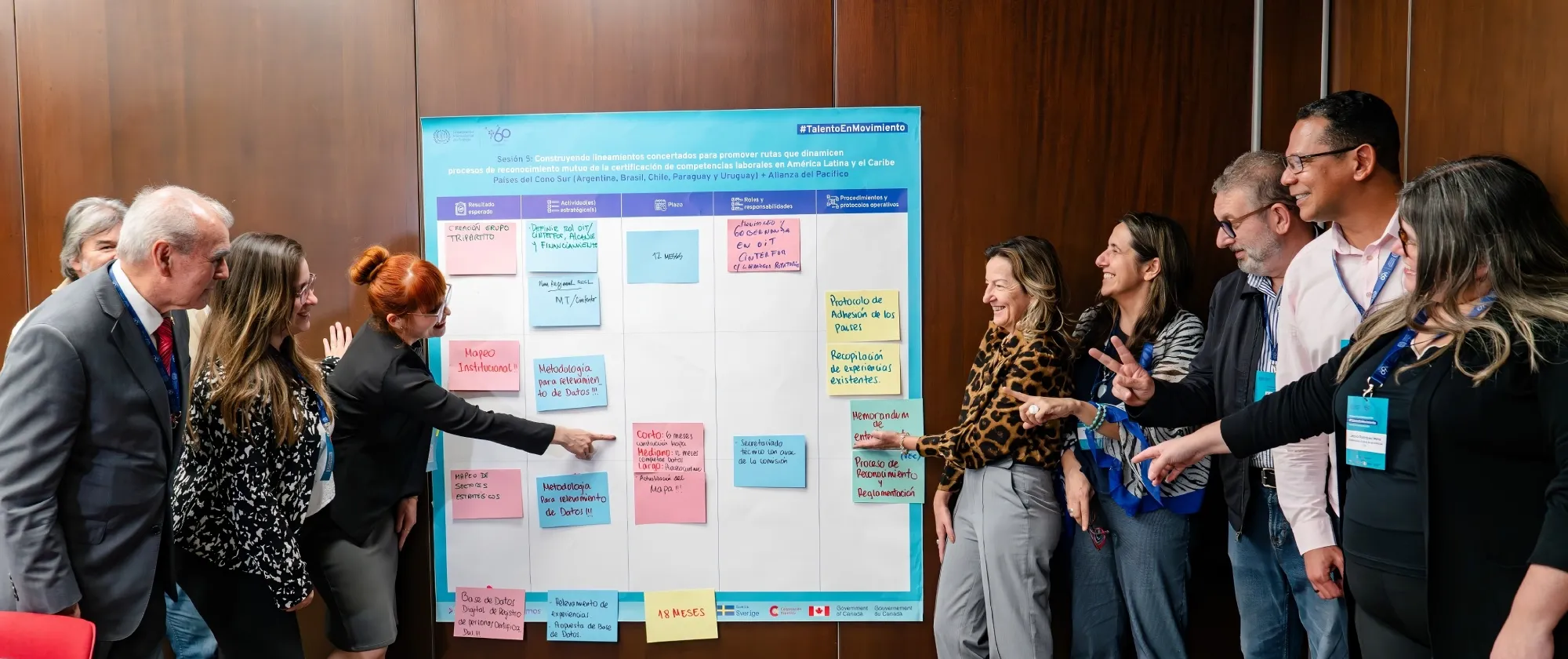 Grupo de nueve personas adultas de pie frente a un rotafolio con post-its de colores, señalando y comentando las notas durante un taller.