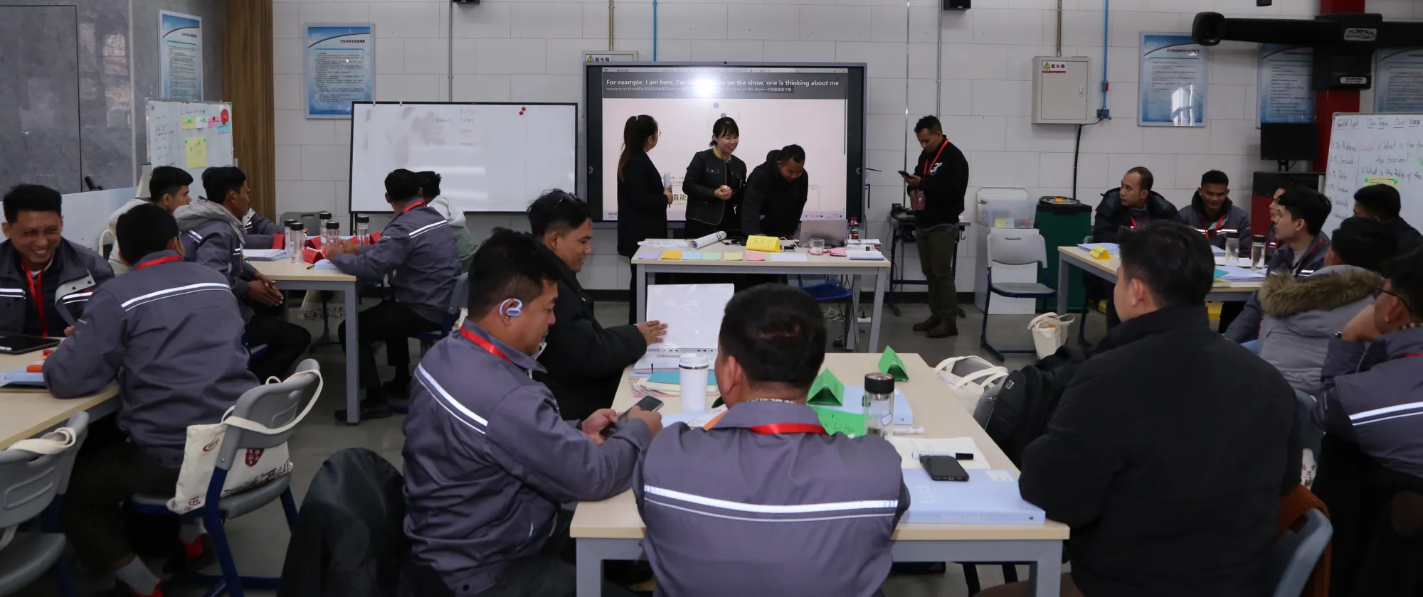 People in uniforms are seated, while a few individuals stand at the front of the class near a whiteboard.
