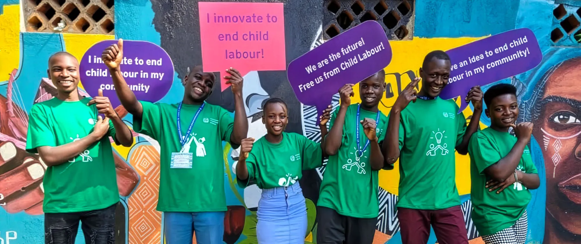 A group of young people in green T-shirts stand in front of a colourful mural, smiling and holding signs calling for innovations to end child labour in their communities.