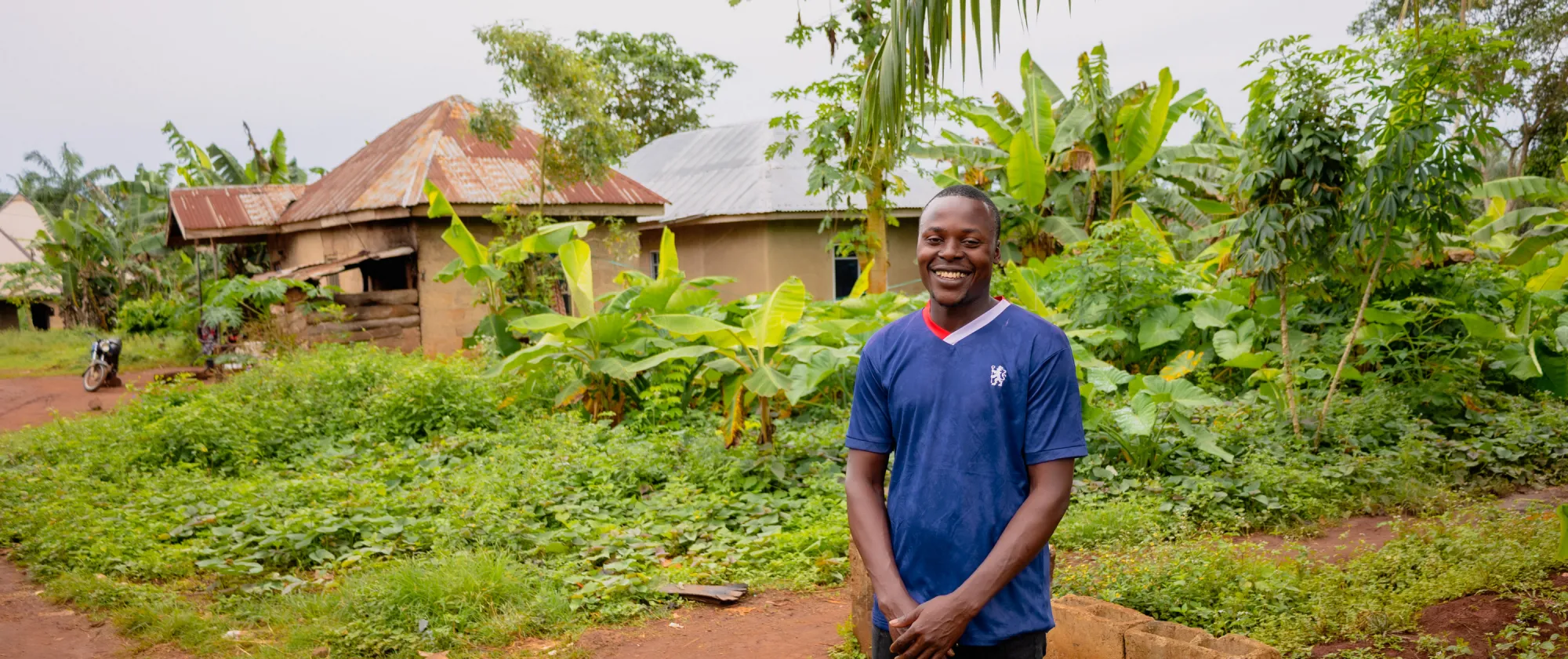 A young Nigerian man, David Chukwuemeka, stands smiling in front of his family’s rural home in Aponmu, Ondo State. He is wearing a blue shirt, surrounded by green plants and simple houses in the background.