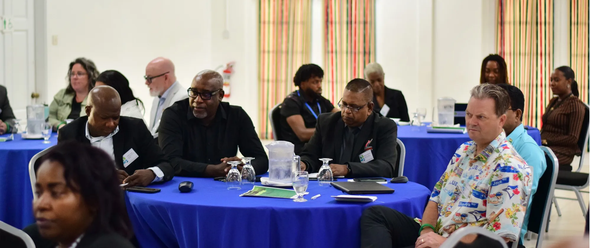 Participants sit at round tables covered in blue cloths during a formal meeting, listening attentively to proceedings at the front of the room. 