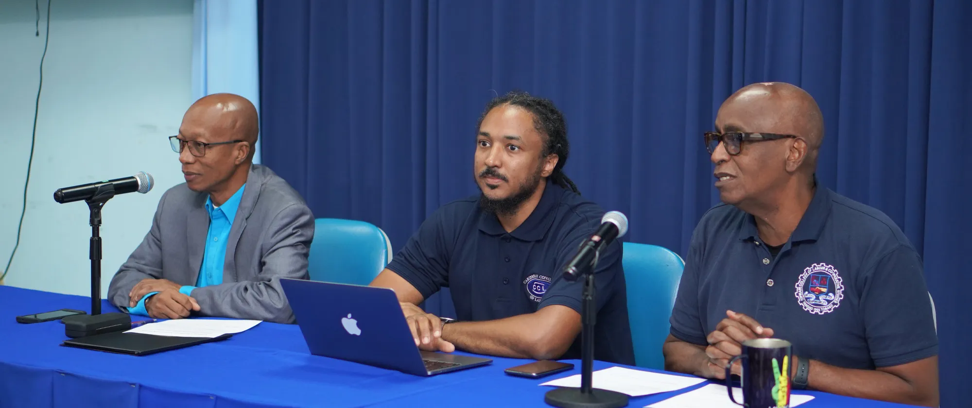 Three men sit side by side at a blue-covered table in front of a blue curtain, each with a microphone and documents in front of them.