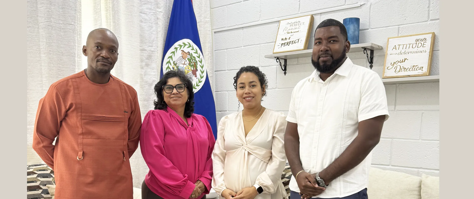 A group portrait of four people standing indoors in front of a Belize flag, smiling at the camera