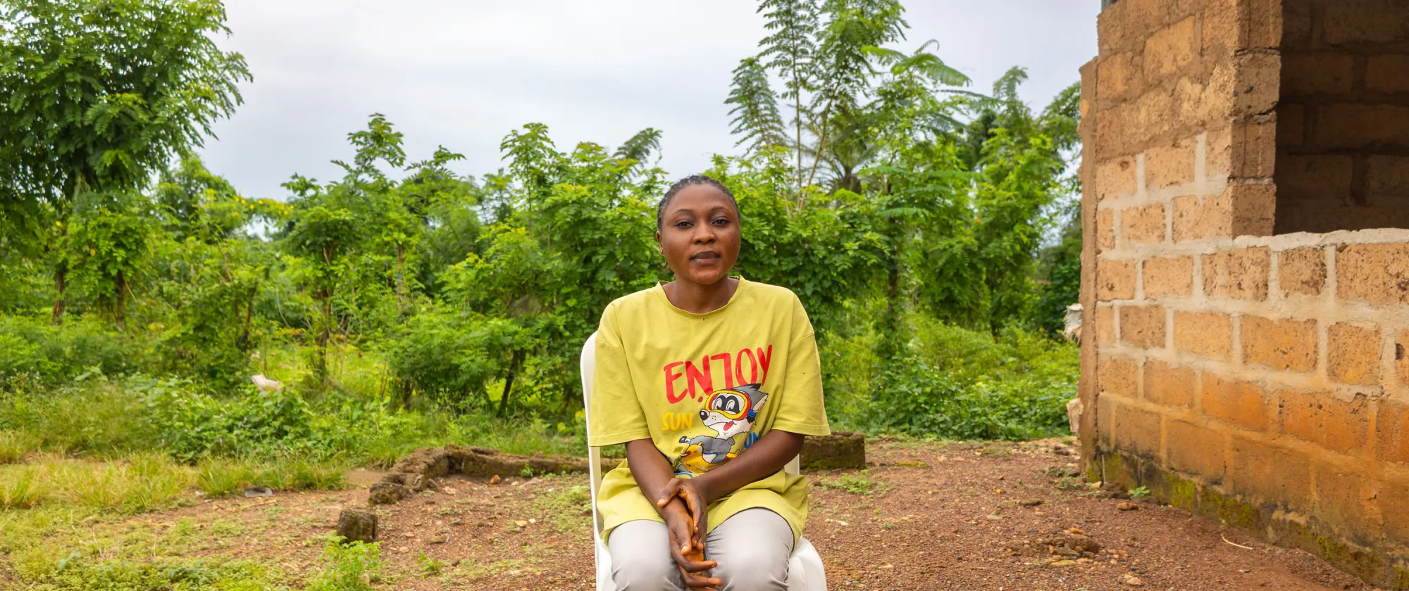 A young woman, Oyefunde Bukola, sits on a white plastic chair outside her home in rural Ondo State, Nigeria. She faces the camera calmly, surrounded by lush green trees and a partially built brick structure.