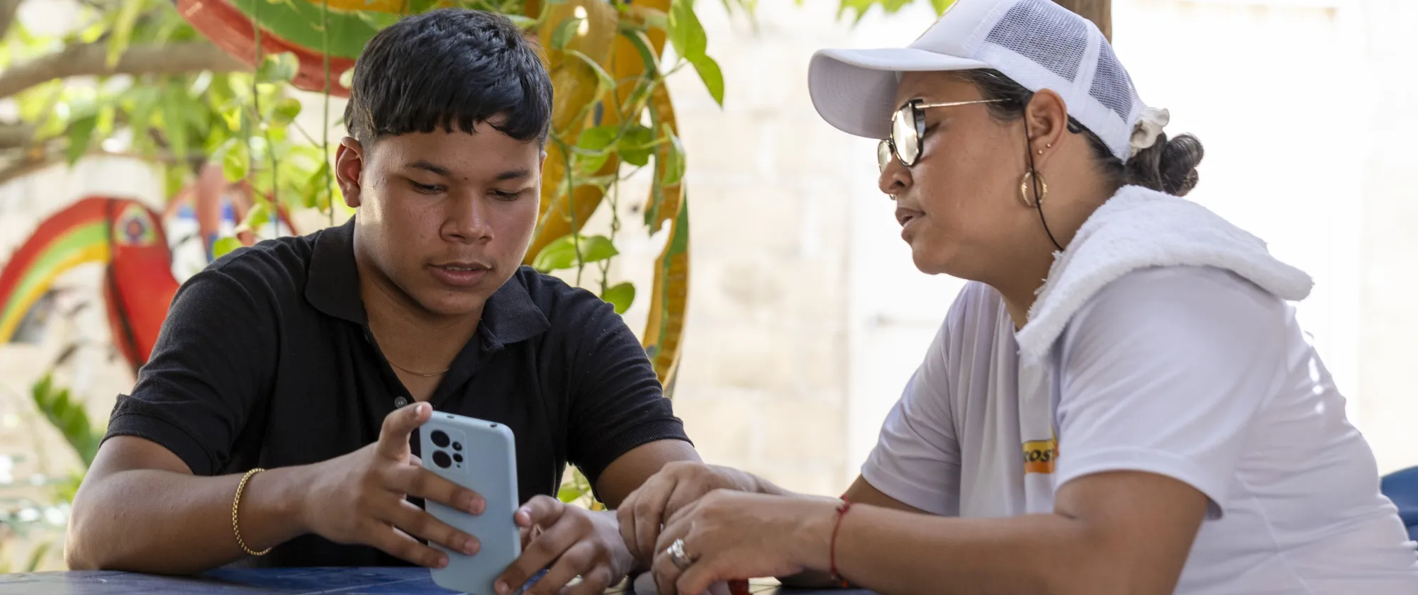 Rafael Meza y su mamá, Marlin Judith, están sentados juntos a una mesa. Rafael le está mostrando su teléfono móvil.