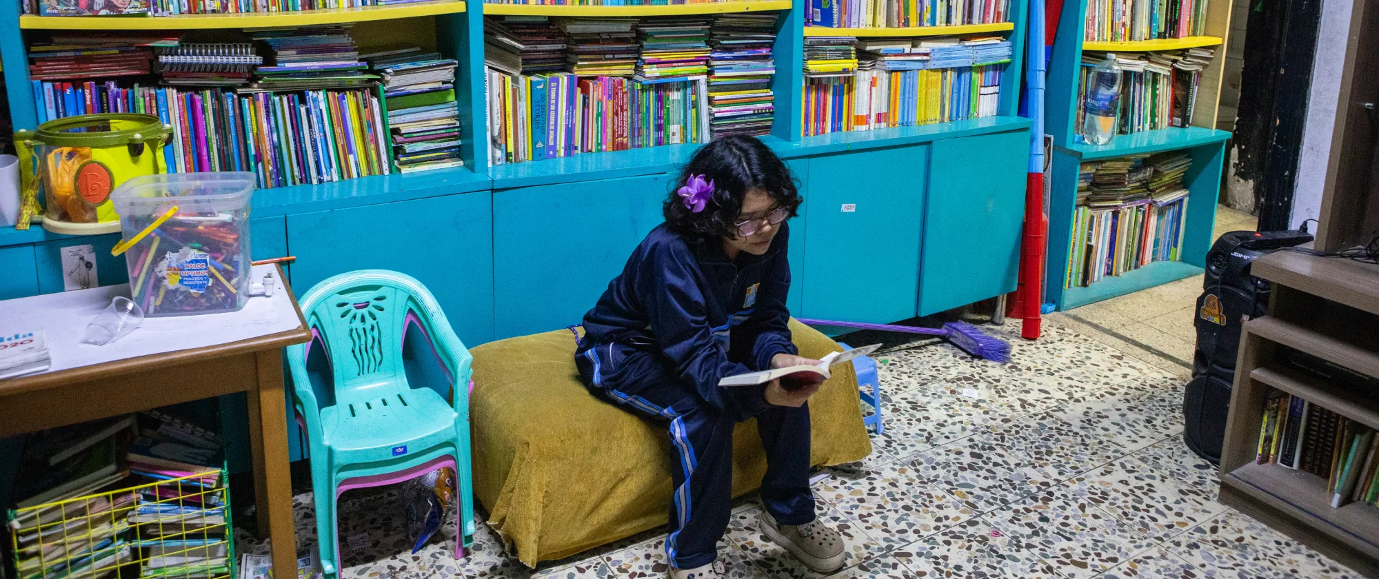 A teenage girl sits on a small bench inside a neighbourhood library, reading a book. Shelves filled with books line the wall behind her.